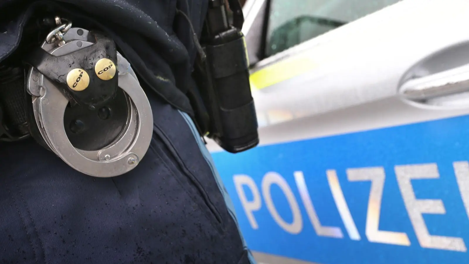 Ein Polizist mit Handschellen und einer Pistole am Gürtel steht vor einem Streifenwagen. In Oberbayern hat ein Seniorenpaar nun eine Betrügerbande hereingelegt und einen der Täter festnehmen lassen. (Archivbild-Illustration) (Foto: Karl-Josef Hildenbrand/dpa)