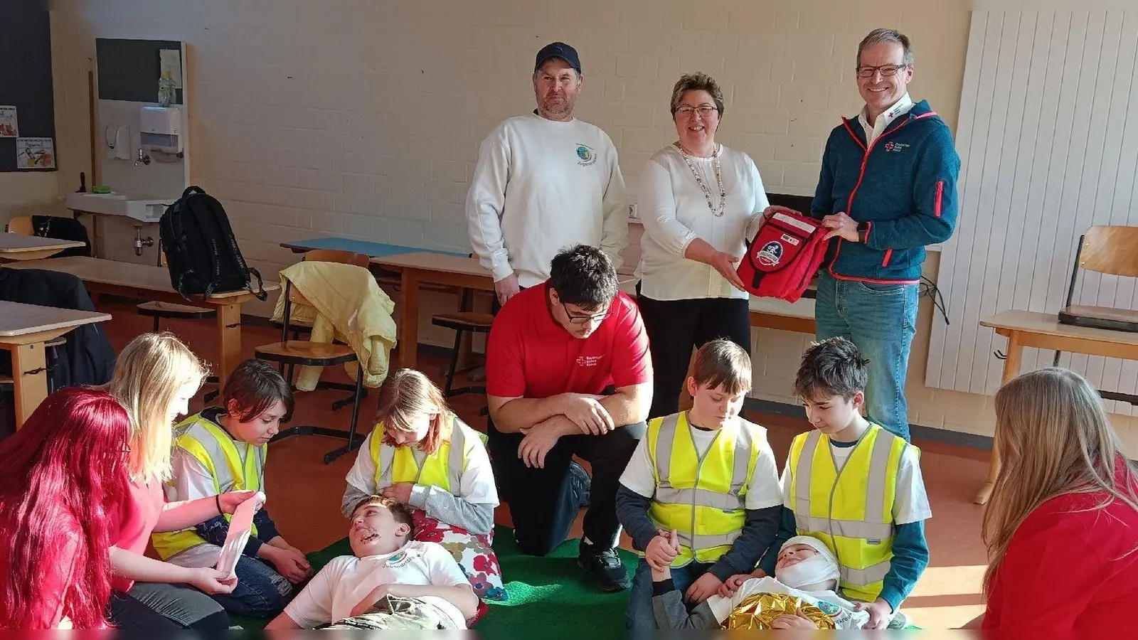 Die Uehlfelder Schülerinnen und Schüler Mandy Wick, Lena Kutzner, Julian Negele, Sulejman Kadiric (liegend), Cecilia Weimann, Dominik Wehrling, Hannes Detzel, Aaron Grimm, Syla Hasanaj (liegend) und Michelle Kirchhöfer (von links) haben eine Rettungsszene nachgestellt, während BRK-Kreisgeschäftsführer Ralph Engelbrecht Silke Schür und Uwe Ficht (hinten von rechts) den Rettungsrucksack überreicht. (Foto: Uta Besendörfer)