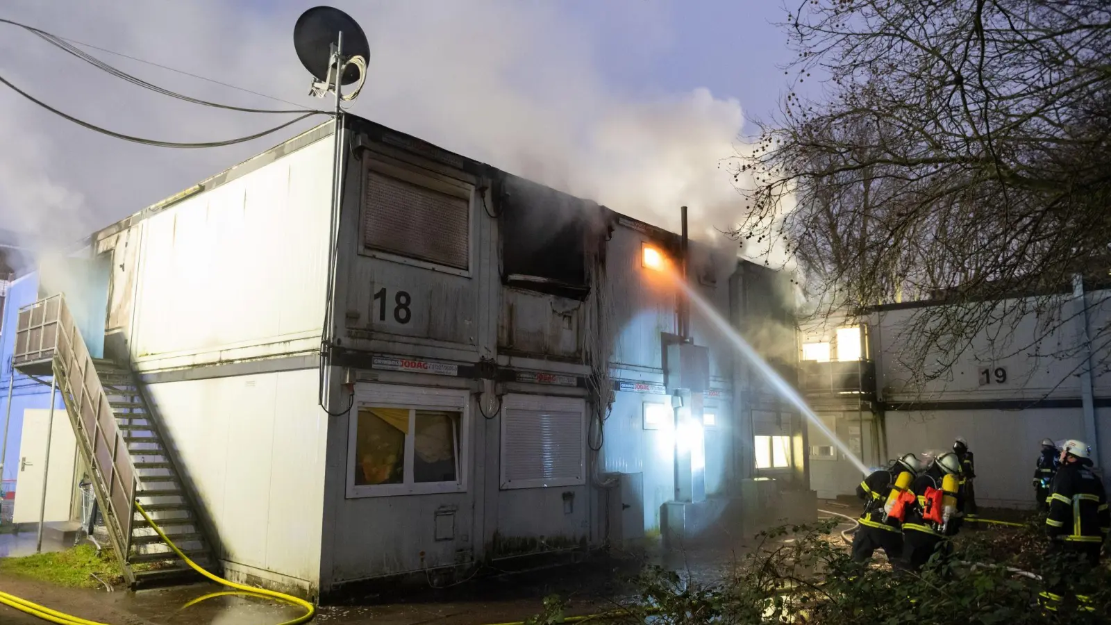 Feuerwehrleute stehen bei Löscharbeiten vor den verqualmten und teils brennenden Container-Einheiten einer Flüchtlingsunterkunft in Hamburg-Winterhude. (Foto: Jonas Walzberg/dpa)