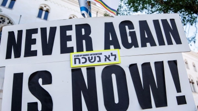 Ein Schild mit der Aufschrift „Never again is now!“ ist bei Teilnehmern einer Mahnwache gegen ein geplantes Pro-Palästina-Protestcamp vor der Ludwig-Maximilians-Universität (LMU) zu sehen. (Foto: Matthias Balk/dpa)