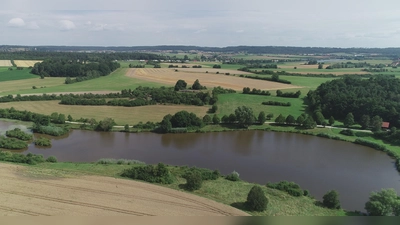 Der Stausee bei Dorfgütingen mit dem Festival-Gelände: Der Zugangsbereich wird nach gegenwärtiger Planung unweit der Fischerhütte (rechts) aufgebaut. Davor ist ein öffentlich zugänglicher Biergarten geplant, an dessen Betrieb örtliche Vereine schon ihr Interesse bekundet haben. Die Grünfläche links von dem Waldstück ist als Campingplatz und Backstage-Bereich vorgesehen. Derweil soll die Bühne bei der Hecke aufgestellt werden, die die beiden Wiesen voneinander abgrenzt. Als Parkplatz ist ein im Hintergrund zu sehendes Areal ins Auge gefasst. (Foto: Stadt Feuchtwangen/Werner Tippmann)