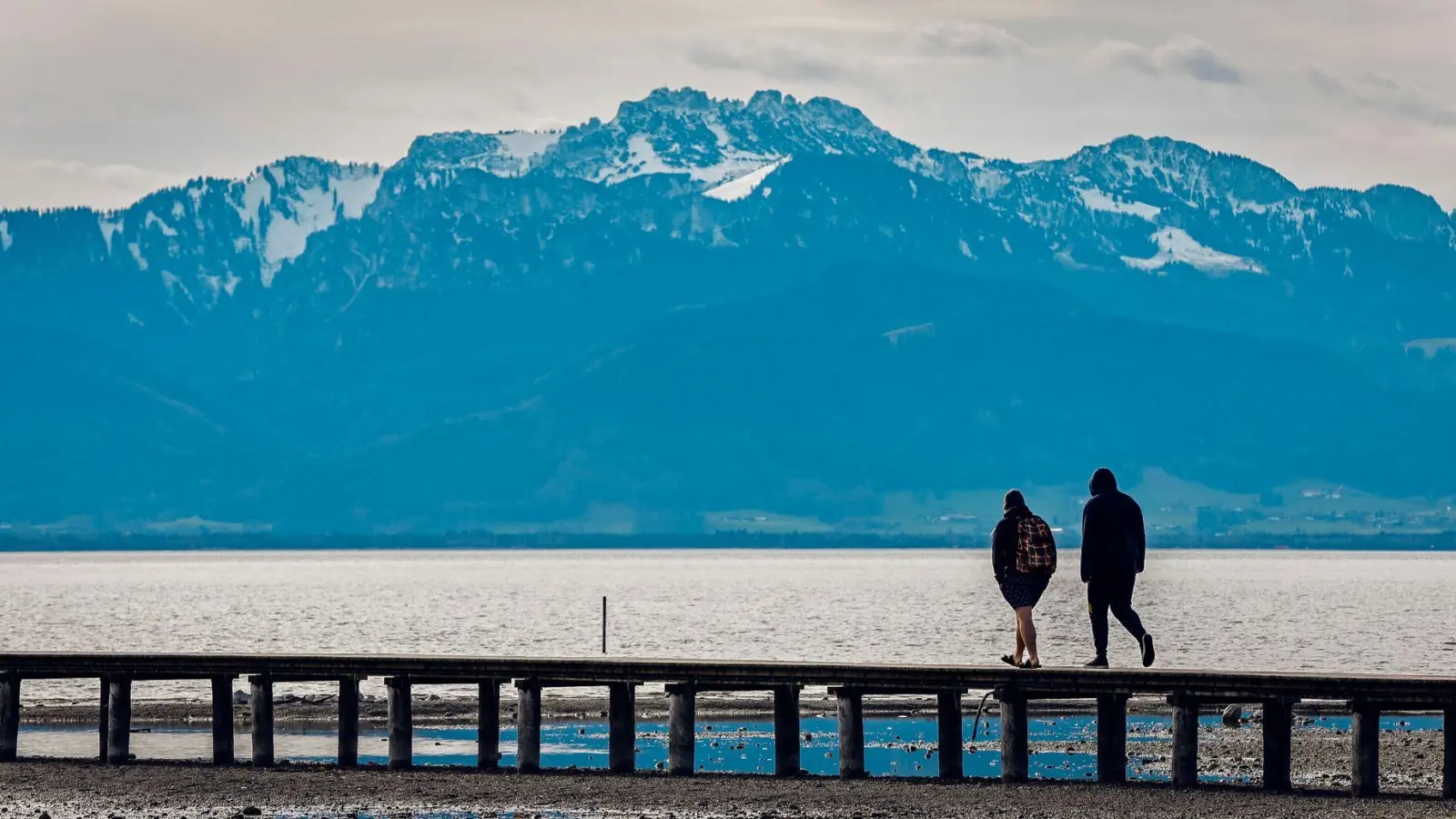 Zwei Männer gehen bei frühlingshaften Temperaturen vor der fast schneefreien Kulisse der Kampenwand in den Chiemgauer Bergen über einen Steg im Chiemsee. (Foto: Uwe Lein/dpa)