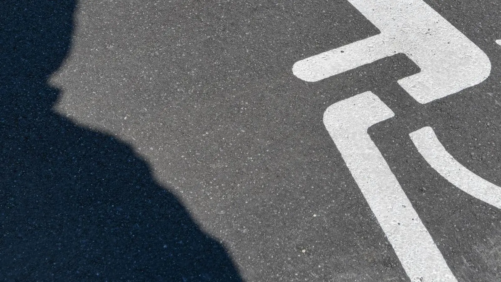 Ein Piktogramm für einen Behindertenparkplatz und ein Schatten einer Person sind auf dem Asphalt zu sehen. (Foto: Patrick Pleul/dpa-Zentralbild/dpa/Archivbild)