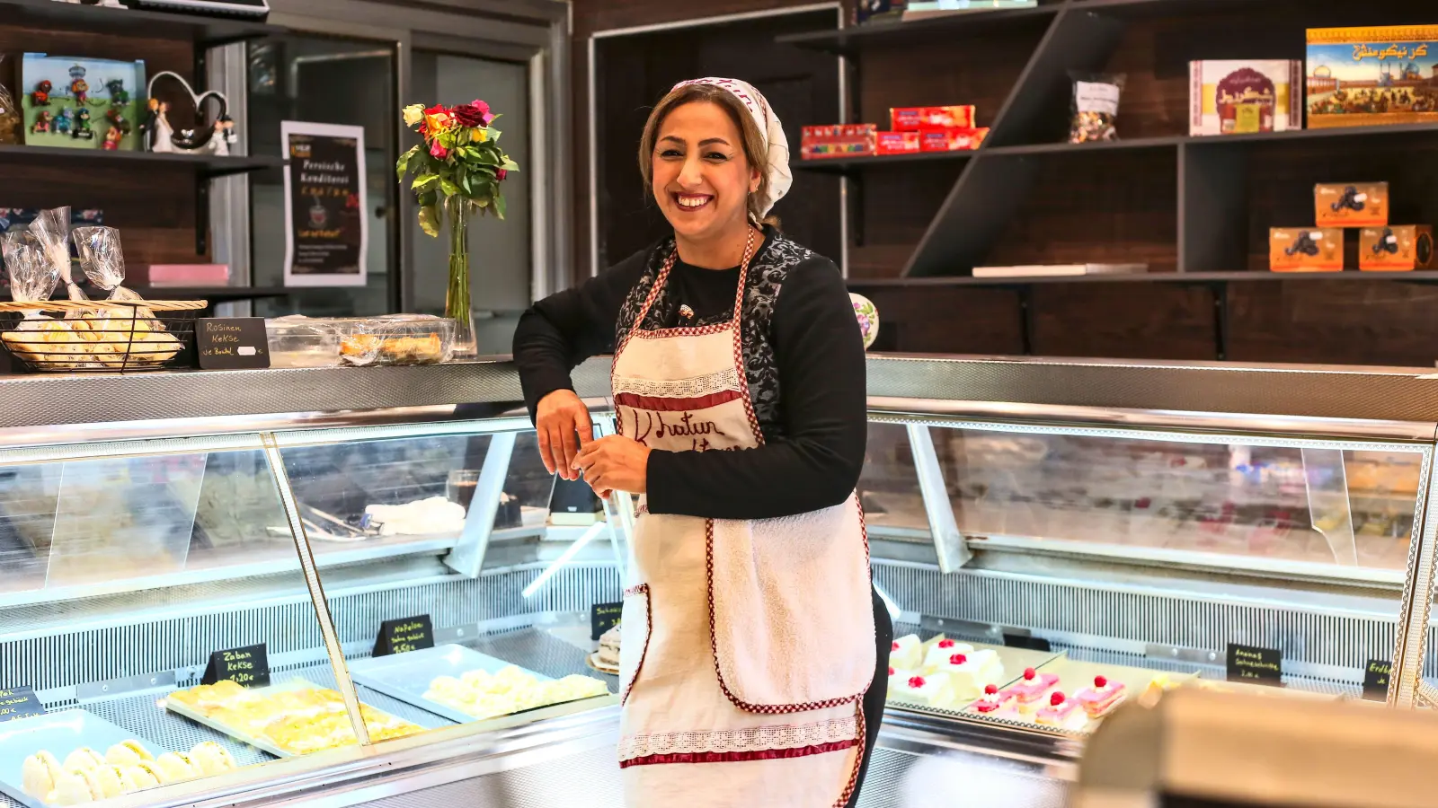 Die Theke der Feinbäckerei ist reichlich mit klassischem Gebäck aus dem Iran und deutschen Torten gefüllt. Davor strahlt die 42-jährige Khadijeh Jamei. (Foto: Tizian Gerbing)