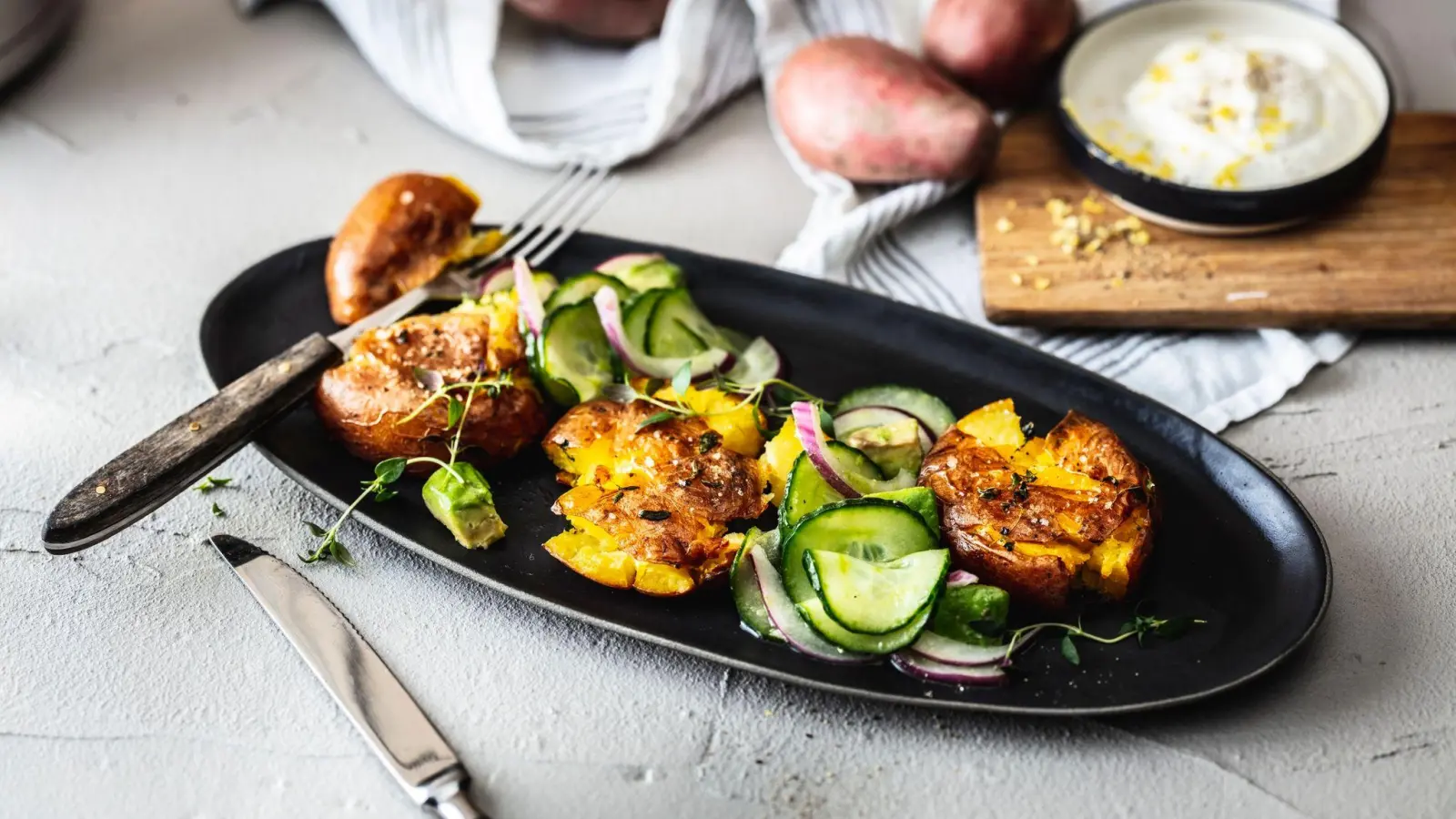 Die knusprig-krossen Smashed Potatoes werden mit Kartoffeln vom Vortag gebacken und mit Gurken-Avocado-Salat und Zitronen-Schmand angerichtet. (Foto: KMG/die-kartoffel.de/dpa-tmn)