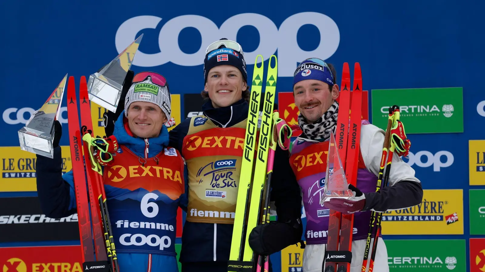 Johannes Hoesflot Klaebo (M) aus Norwegen jubelt neben dem Zweitplatzierten Mika Vermeulen (l) aus Österreich und dem Drittplatzierten Hugo Lapalus aus Frankreich auf dem Podium der Tour de Ski. (Foto: Alessandro Trovati/AP/dpa)